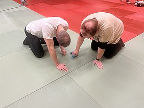 Zwei Personen knien auf einer Tatami-Matte und reinigen den Boden mit Tüchern. Der Raum hat abwechselnd grüne und rote Matten. Diese Aktivität deutet auf eine Vorbereitung oder Reinigung nach einem Training hin.