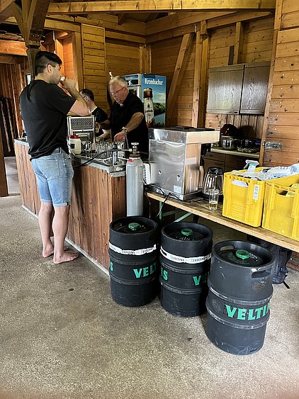 Ein Mann steht an einer Holzbar und bedient sich an einem Getränk. Hinter ihm arbeitet ein weiterer Mann, während Bierfässer mit der Aufschrift "Veltins" am Stadtrand stehen. Der Raum hat eine rustikale Atmosphäre aus Holz.