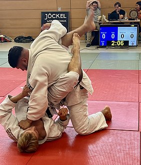 Zwei Judokas in weißen Anzügen kämpfen auf einer roten Matte. Ein Kämpfer hat seinen Gegner in einer Hebelposition, während der andere auf dem Rücken liegt. Im Hintergrund sind Zeitnehmer und Zuschauer sichtbar. Die Wettkampfzeit beträgt noch 2 Minuten und 6 Sekunden.