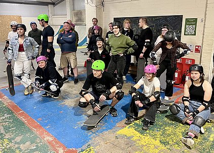 Eine Gruppe von Skateboardern, teilweise in Schutzkleidung und Helmen, versammelt in einer Indoor-Skateanlage. Einige sitzen auf dem Boden mit Skateboards, während andere stehen. Die Stimmung ist entspannt und die Teilnehmer scheinen sich auf eine Skate-Session vorzubereiten.