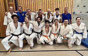 Eine Gruppe von 14 Männern in weißen Judogi steht und sitzt zusammen auf einer Judomatte. Einige tragen farbige Gürtel, darunter schwarz, orange und blau. Im Hintergrund ist eine Holzwand sichtbar. Die Stimmung ist freundlich und die Männer lächeln.