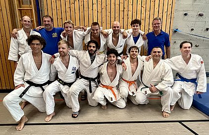 Eine Gruppe von 14 Männern in weißen Judogi steht und sitzt zusammen auf einer Judomatte. Einige tragen farbige Gürtel, darunter schwarz, orange und blau. Im Hintergrund ist eine Holzwand sichtbar. Die Stimmung ist freundlich und die Männer lächeln.