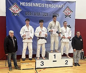 Die Hessenmeisterschaft U15/U18/U21 2025 zeigt vier Judoka auf einem Siegerpodest. Die Athleten halten Urkunden in den Händen. Der Erste trägt eine Medaille. Neben ihnen stehen zwei Trainer oder Offizielle, die stolz die Leistung der Sportler würdigen. Im Hintergrund ist ein Banner mit dem Turniernamen.