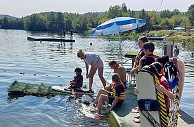 Eine Gruppe von Kindern in Schwimmwesten steht am Ufer eines Badegewässers. Ein Junge sitzt auf einem Surfbrett im Wasser, während eine Frau ihm hilft. Im Hintergrund sind weitere Personen auf einem Ponton und im Wasser zu sehen. Der Tag ist sonnig und die Umgebung ist grün.