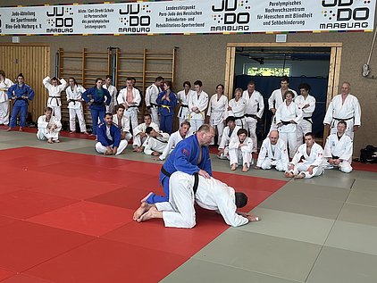 Eine Gruppe von Judo-Praktizierenden in einem Dojo. Im Vordergrund demonstriert ein Trainer eine Technik an einem Teilnehmer, während andere in Judokleidung zuschauen. Die Atmosphäre ist konzentriert und lehrreich, mit roten und grünen Matten im Hintergrund.