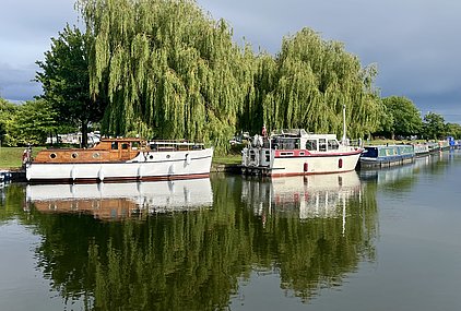 Zwei Boote liegen am Ufer eines ruhigen Kanals, umgeben von üppigem Grün und Weidenbäumen. Die Boote spiegeln sich im Wasser, während der Himmel leicht bewölkt ist. Die Szene vermittelt eine friedliche, naturnahe Atmosphäre.