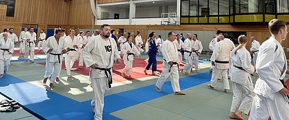 Eine Gruppe von Judokas in weißen und blauen Anzügen trainiert auf einer Kampffläche. Die Teilnehmer bewegen sich in verschiedenen Richtungen und tragen unterschiedliche Gürtel, darunter schwarze und braune. Die Atmosphäre ist dynamisch und sportlich.