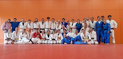Eine Gruppe von etwa 30 Menschen in Judokleidung steht in mehreren Reihen auf einer Matte. Sie lächeln und posieren, während eine orangene Wand im Hintergrund sichtbar ist. Einige tragen verschiedene Gürtel, die ihre Graduierung im Judo anzeigen.