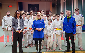 Eine Gruppe von 18 Judoka, darunter Kinder und Erwachsene, stellt sich in einem Trainingsraum auf. Im Vordergrund stehen zwei Frauen und zwei Männer, die lächeln. Die Judoka tragen weiße Gürtel und einige haben orange Gürtel. Der Boden ist mit matten Abdeckung versehen.