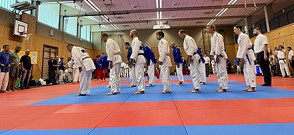 Eine Gruppe von sieben Judokas in weißen und blauen Judokis steht in einer Reihe auf einer Mattenfläche. Sie beugen sich leicht nach vorne, möglicherweise als Teil einer Zeremonie oder Begrüßung. Im Hintergrund sind Zuschauer und weitere Teilnehmer zu sehen.