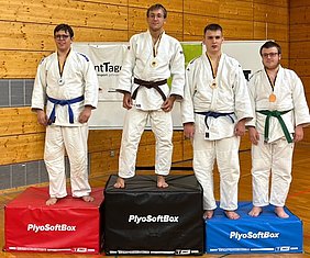 Vier Judo-Kämpfer stehen auf Treppchen aus Softboxen, die mit verschiedenen Farben gekennzeichnet sind. Der erste Platz (Mitte) trägt eine goldene Medaille, der zweite Platz eine silberne, der dritte Platz eine bronzene. Alle tragen Judogis und zeigen stolz ihre Auszeichnungen.
