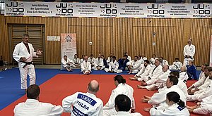 Ein Judo-Trainer steht in einem Trainingsraum und erklärt etwas vor einer Gruppe von Judo-Praktizierenden, die auf einer blauen und roten Matte sitzen. Im Hintergrund sind Banner und Holzwände sichtbar. Die Teilnehmer tragen traditionelle Judo-Uniformen.