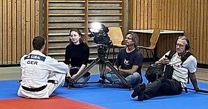 Ein Judoka mit dem Namen D. Goral mit einer weißen Judojacke sitzt auf einer blauen Matte und spricht mit einer Person, die ihm gegenüber sitzt. Im Hintergrund sind zwei Kameramänner und ein weiterer Mann, der ein Aufnahmegerät benutzt.
