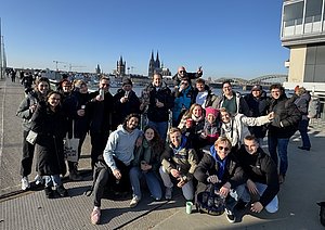Eine Gruppe von etwa 20 Personen steht am Ufer in Köln, lächelt und posiert für das Foto. Im Hintergrund sind die Kölner Domtürme und eine Brücke sichtbar. Die Sonneneinstrahlung sorgt für eine helle und freundliche Atmosphäre.