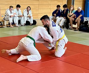 Zwei Judokas im Wettkampf auf einer roten Matte. Der Kämpfer mit dem grünen Gürtel versucht, einen Griff am Gegner mit dem gelben Gürtel zu bekommen. Im Hintergrund sitzen Zuschauer, die das Geschehen beobachten.