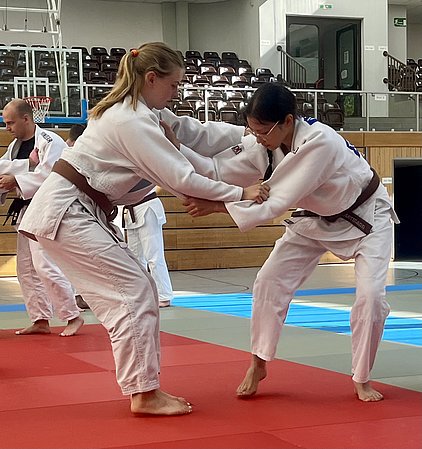 Zwei Judokas in Ippon-Position üben miteinander auf einer roten Matte in einem Sporttraining. Beide tragen Judopas mit braunen Gurten. Im Hintergrund sind weitere Teilnehmer sichtbar, die ebenfalls trainieren.