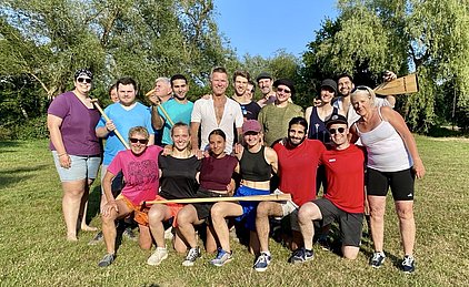 Eine Gruppe von 18 Personen steht auf einem grünen Feld. Sie lächeln und posieren gemeinsam, einige halten Paddel in den Händen. Die Atmosphäre wirkt freundschaftlich und gesellig, umgeben von Bäumen und blauem Himmel. Es scheinen aktive Freizeit- oder Sportler zu sein.
