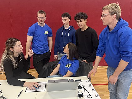Eine Frau sitzt an einem Tisch und spricht mit einer Gruppe von fünf jungen Männern, die um sie herum stehen. Die Männer tragen unterschiedliche sportliche Oberteile. Auf dem Tisch befinden sich ein Laptop und einige Unterlagen. Der Hintergrund ist rot.