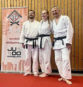 Drei Personen in weißen Judo-Anzügen mit schwarzen Gürteln stehen auf einer roten Matte. Sie lächeln und posieren für das Foto. Im Hintergrund ist ein Banner mit dem Text „Herzlich Willkommen“ und dem Logo des Judoverbands zu sehen.