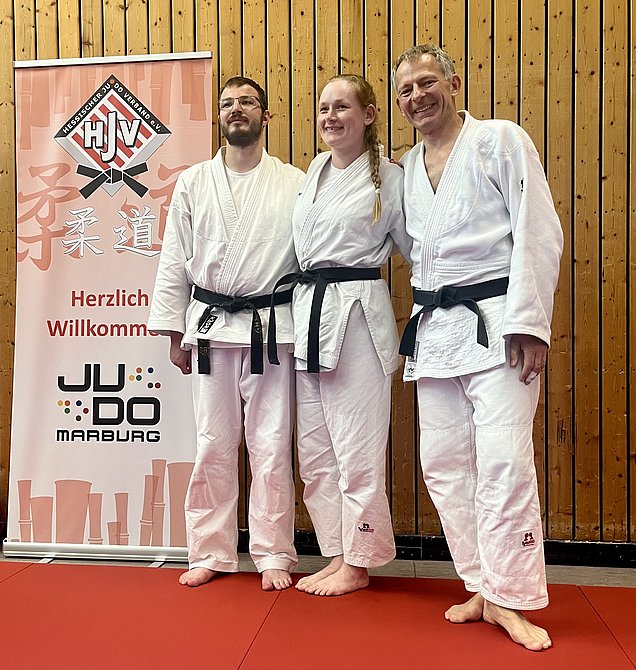 Drei Personen in weißen Judo-Anzügen mit schwarzen Gürteln stehen auf einer roten Matte. Sie lächeln und posieren für das Foto. Im Hintergrund ist ein Banner mit dem Text „Herzlich Willkommen“ und dem Logo des Judoverbands zu sehen.