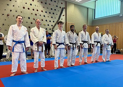 Sechs Judokas stehen in einer Reihe auf einer Judomatte, jeder in einem weißen Gi, einige mit farbigen Gürteln. Sie sind bereit für eine Veranstaltung, im Hintergrund sind Zuschauer zu sehen. Die Atmosphäre ist sportlich und konzentriert.