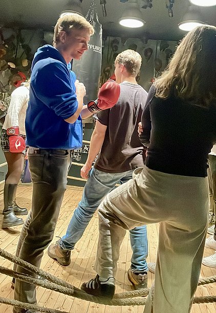Eine Gruppe junger Menschen steht in einem Boxraum. Ein Junge in einem blauen Hoodie hält einen roten Boxhandschuh. Im Hintergrund sind weitere Personen zu sehen, einige tragen auch Boxhandschuhe. Der Raum ist mit Boxutensilien dekoriert und hat Holzboden.