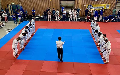 Eine Gruppe von Judoka in weißen Anzügen steht in zwei Reihen auf blauen und roten Matten. Ein Trainer in einem weißen Hemd steht in der Mitte und zeigt mit den Händen Anweisungen. Zuschauer beobachten die Szene im Hintergrund.