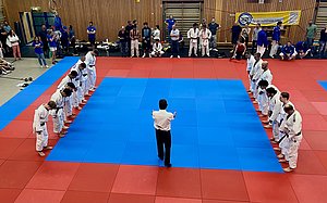 Eine Gruppe von Judoka in weißen Anzügen steht in zwei Reihen auf blauen und roten Matten. Ein Trainer in einem weißen Hemd steht in der Mitte und zeigt mit den Händen Anweisungen. Zuschauer beobachten die Szene im Hintergrund.
