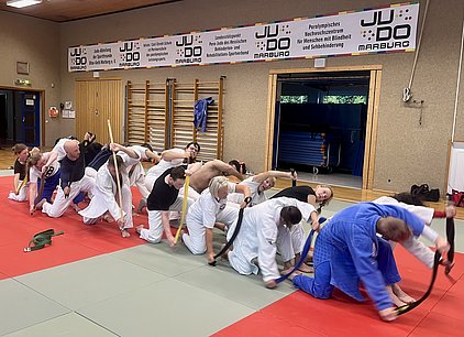 Eine Gruppe von Judokas in Trainingsanzügen übt auf einer Judomatte. Einige Teilnehmer halten Trainingsgeräte und bewegen sich synchron. Die Atmosphäre ist konzentriert und dynamisch, während die Sportler ihre Techniken in einer Judohalle verbessern. Im Hintergrund sind Wände mit Informationsplakaten sichtbar.
