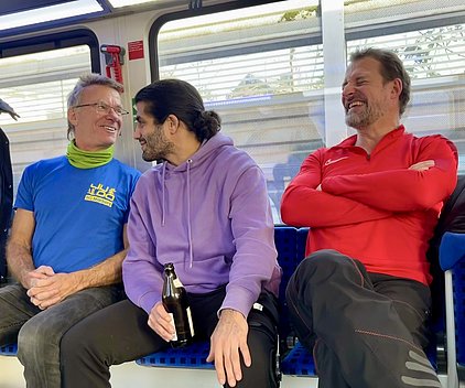 Drei Männer sitzen in einer Bahn, lachen und unterhalten sich entspannt. Der Mann links trägt ein blaues Shirt mit grünem Schal, der in der Mitte hat einen lilafarbenen Pullover und hält eine Flasche, der Mann rechts hat ein rotes Shirt und verschränkt die Arme.