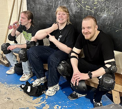 Drei Personen sitzen auf einer Bank in Schutzkleidung für Sportarten wie Skateboarding. Eine Person trinkt aus einer Flasche, während eine andere lächelt und auf ihren Arm zeigt. Die Stimmung ist entspannt und freundlich, mit einer Tafel im Hintergrund, auf der Kritzeleien zu sehen sind.