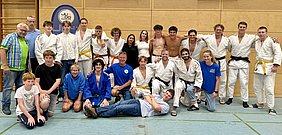 Eine Gruppe von 22 Personen steht zusammen in einem Raum, in Sportbekleidung, überwiegend in weißen Judogi. Einige tragen Gürtelfarben in Gelb und Schwarz. Die Szenerie wirkt freundlich, mit lächelnden Gesichtern, und es gibt ein Wappen des Judo-Vereins im Hintergrund.