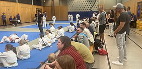 Das Bild zeigt eine Sporthalle mit einer Gruppe von Kindern in weißen und orangefarbenen Judoanzügen, die auf Matten sitzen. Im Hintergrund sind Zuschauer und Trainer zu sehen, die das Geschehen beobachten. Einige Kinder trainieren und andere warten auf ihren Einsatz.