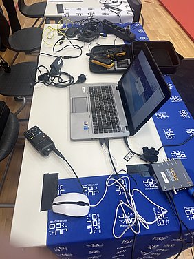 Auf einem Tisch liegen ein Laptop, eine Maus, ein Funkgerät und mehrere Kabel. Der Laptop zeigt eine Benutzeroberfläche. Daneben befindet sich ein Werkzeugkoffer sowie eine Steckdosenleiste. Der Tisch ist mit einer blauen Tischdecke bedekt, die mit einem Judo-Logo verziert ist.