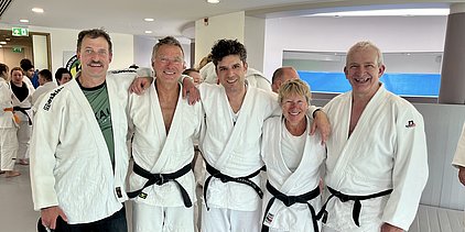 Fünf Personen in weißen Judogis und schwarzen Gürteln stehen lächelnd nebeneinander. Sie sind in einem modernen Raum mit anderen Judokas im Hintergrund. Alle wirken freundlich und entspannt, was eine Atmosphäre der Gemeinschaft und des Trainings vermittelt.
