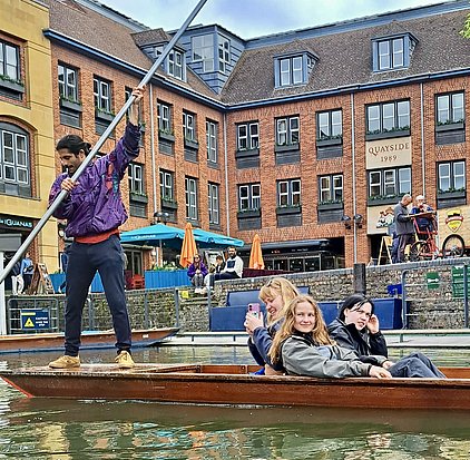 Vier Personen sitzen in einem Boot auf einem Kanal. Ein Mann steht am Bug und steuert mit einem langen Stock. Eine Frau hält ein Handy und lächelt. Im Hintergrund sind Gebäude und weitere Menschen zu sehen.