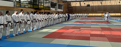 Eine Gruppe von Judokas in weißen Gi steht in zwei Reihen auf einer Judomatte. Einige tragen schwarze Gürtel, während andere blauen Gürtel tragen. Die Halle ist gut beleuchtet und bietet eine offene, sportliche Atmosphäre.