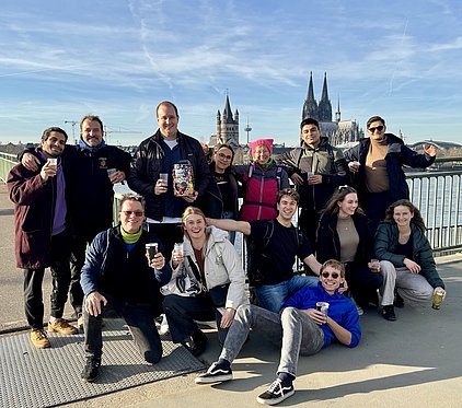 Eine Gruppe von zwölf Menschen posiert fröhlich an einer Brücke mit Blick auf die Kölner Skyline. Einige halten Getränke in der Hand. Die Stimmung ist entspannt und festlich, umgeben von klaren Himmel und Wasser im Hintergrund.