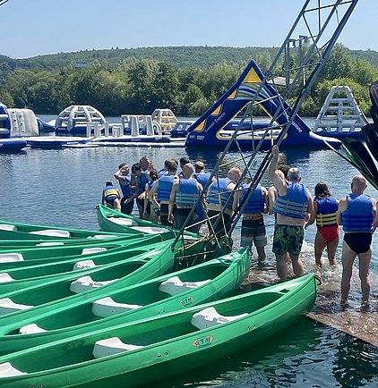 Grüne Kanus liegen am Ufer eines Gewässers. Eine Gruppe von Menschen in Schwimmwesten steht am Wasser und bereitet sich darauf vor, ins Wasser zu gehen. Im Hintergrund sind schwimmende Wasserattraktionen sichtbar.