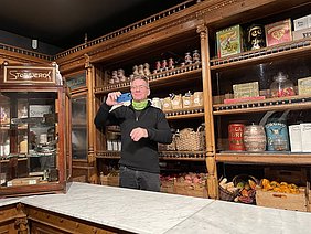 Ein Mann steht an einem Marmortisch und hält ein Smartphone in der Hand, während er sich selbst filmt. Im Hintergrund ist ein nostalgisches Regal mit verschiedenen Lebensmitteln und Produkten, darunter Kakaodosen und Körbe mit Obst und Gemüse.