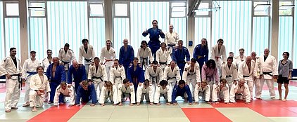 Eine Gruppe von Judokas in Trainingsanzügen steht auf einer Judomatte. Sie bilden eine Menschenpyramide. Die Beteiligten lächeln und zeigen Freude. Der Hintergrund ist hell mit vertikalen Streifen. Die Bildatmosphäre vermittelt Teamgeist und Gemeinschaft.