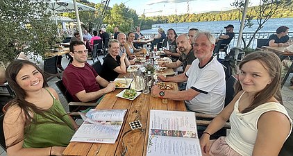 Eine Gruppe von zehn Personen sitzt an einem Tisch im Freien, umgeben von Bäumen und mit Blick auf ein Gewässer. Sie genießen Essen und Getränke, während sie miteinander sprechen. Auf dem Tisch liegen Menüs und verschiedene Speisen.