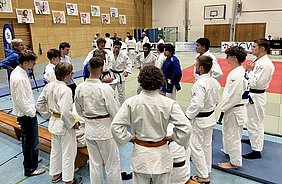 Eine Gruppe von Judokas in traditionellen Judogis versammelt sich in einer Turnhalle. Einige stehen im Kreis und hören einem Trainer zu, während andere auf einer Bank sitzen. Der Hintergrund zeigt Matten und Plakate an Wänden, die mit Judo in Verbindung stehen.