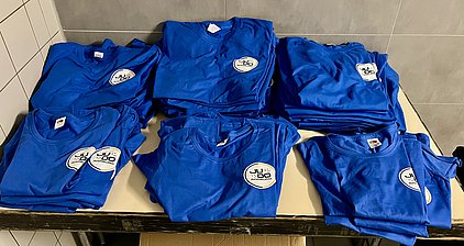Auf dem Bild sind mehrere blaue T-Shirts gestapelt, die ein Logo mit der Aufschrift "Judo Marburg" tragen. Die Shirts liegen ordentlich auf einem Tisch, und der Hintergrund zeigt Fliesen.