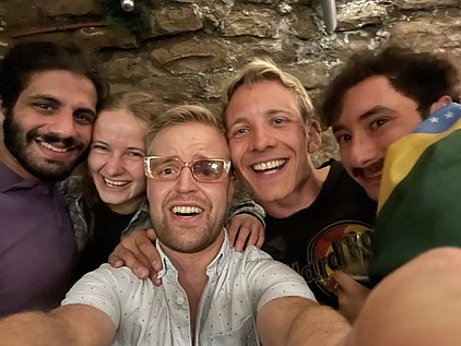 Fünf Personen posieren lächelnd für ein Selfie in einem gemütlichen Raum mit Steinmauern. Sie stehen eng beieinander, wirken freundlich und fröhlich. Einer der Männer hält eine Flagge in den Händen.
