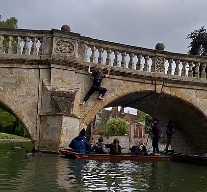Ein Jugendlicher klettert an einer Steinbrücke entlang, während eine Gruppe von Leuten in einem Boot darunter sitzt. Die Szene spielt sich an einem ruhigen Gewässer ab, umgeben von Bäumen und Häusern im Hintergrund. Der Himmel ist bewölkt.
