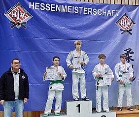 Ein Judo-Wettbewerb während der Hessenmeisterschaft. Vier Kinder stehen auf dem Siegerpodest, jeder hält eine Urkunde. Der zweite Platz wird von einem Jungen mit einer grünen Gürtelfarbe belegt. Ein Erwachsener steht links im Bild. Im Hintergrund ist ein Banner mit dem Schriftzug "HESSENMEISTERSCHAFT".