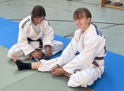 Zwei junge Frauen in Judo-Anzügen sitzen auf Matten in einem Judo-Training. Eine von ihnen lächelt, während die andere an ihren Bändern arbeitet. Die Umgebung deutet auf eine Sporthalle hin.