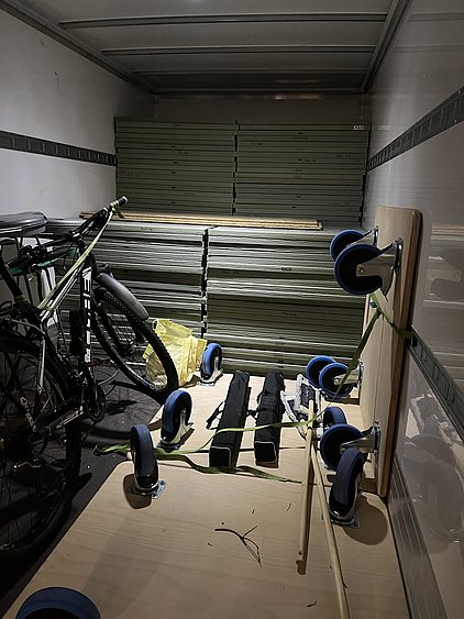 Der Laderaum eines Fahrzeugs ist mit mehreren gestapelten Platten gefüllt. Links steht ein Fahrrad, während ein Holzbrett mit Rollen in der Mitte liegt. Zu sehen sind auch zwei schwarze Objekte und eine gelbe Tüte, die auf dem Boden liegen.
