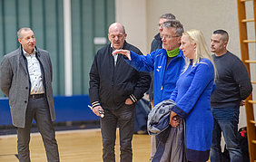Eine Gruppe von Personen steht in einer Sporthalle. Der Mann in blauer Kleidung spricht gestikulierend mit den anderen, darunter Frauen und Männer in unterschiedlichen Outfits. Die Atmosphäre wirkt diskussionsfreudig und aufmerksam.